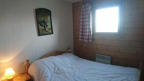 A bedroom with a wooden headboard, a bed with white bedding, a wall-mounted poster, a window with plaid curtains, and a bedside lamp.