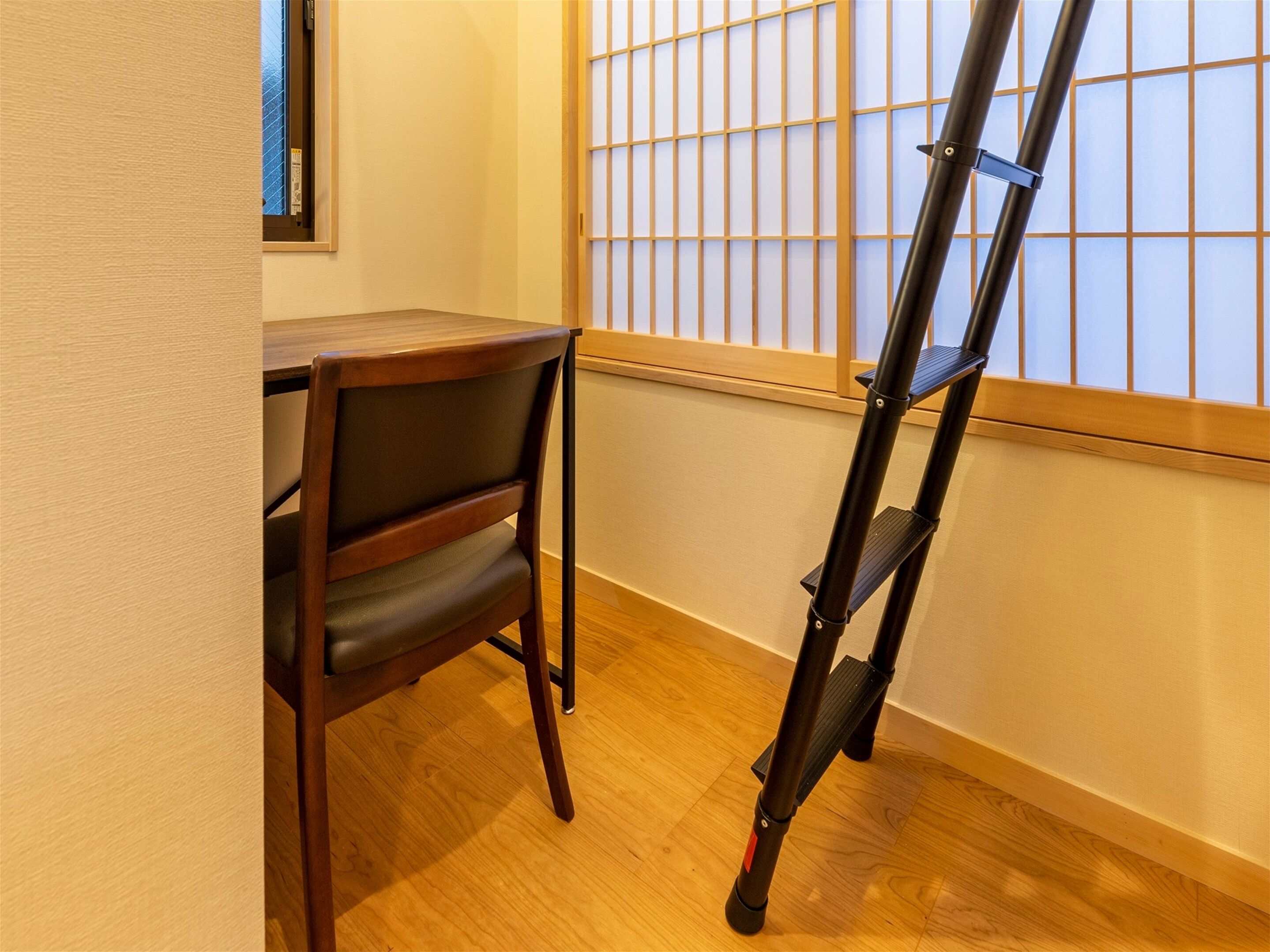 A room with a wooden desk, a chair, and a black folding ladder leaning against the wall.