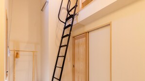 Una habitación con una puerta de madera, una escalera de metal negro apoyada en la pared y un ventanal pequeño.