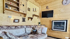 L’intérieur d’un chalet en bois chaleureux et accueillant, avec un canapé, un ours en peluche, des étagères remplies d’objets, une cheminée et une horloge au mur.