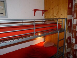 A bunk bed with a red mattress and a pillow, a wooden wall, and a red shelf.