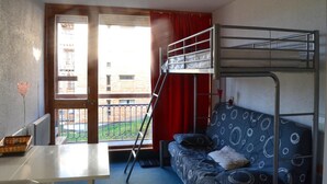 A dormitory room with bunk beds, a sofa, a table, and a window with a view of buildings and greenery. - 100m from ski lifts, 6th floor, balcony, tv, 19m², Les Arcs (Les Arcs)