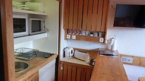 A compact kitchen with a microwave, toaster, and kettle on the counter, and a shelf with mugs and plates above.