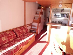 Ein kleiner Raum mit einer Holztreppe, die zu einem Küchenbereich führt, einer Holzbank mit gemustertem Kissen und einer kleinen Uhr an der Wand.