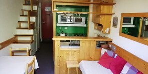 Une pièce compacte avec une kitchenette, un lit agrémenté d’un oreiller rose et un escalier en bois.