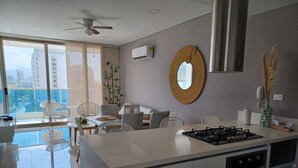 Una cocina moderna con una isla central, un área de comedor con sillas blancas, un ventanal con vistas a edificios y un ventilador de techo.