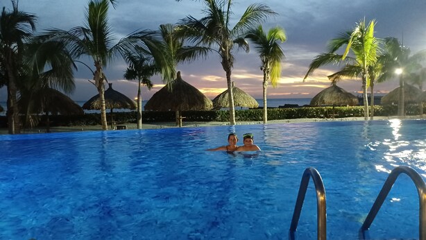 Una piscina con dos personas en el agua, rodeada de palmeras y sombrillas de paja.