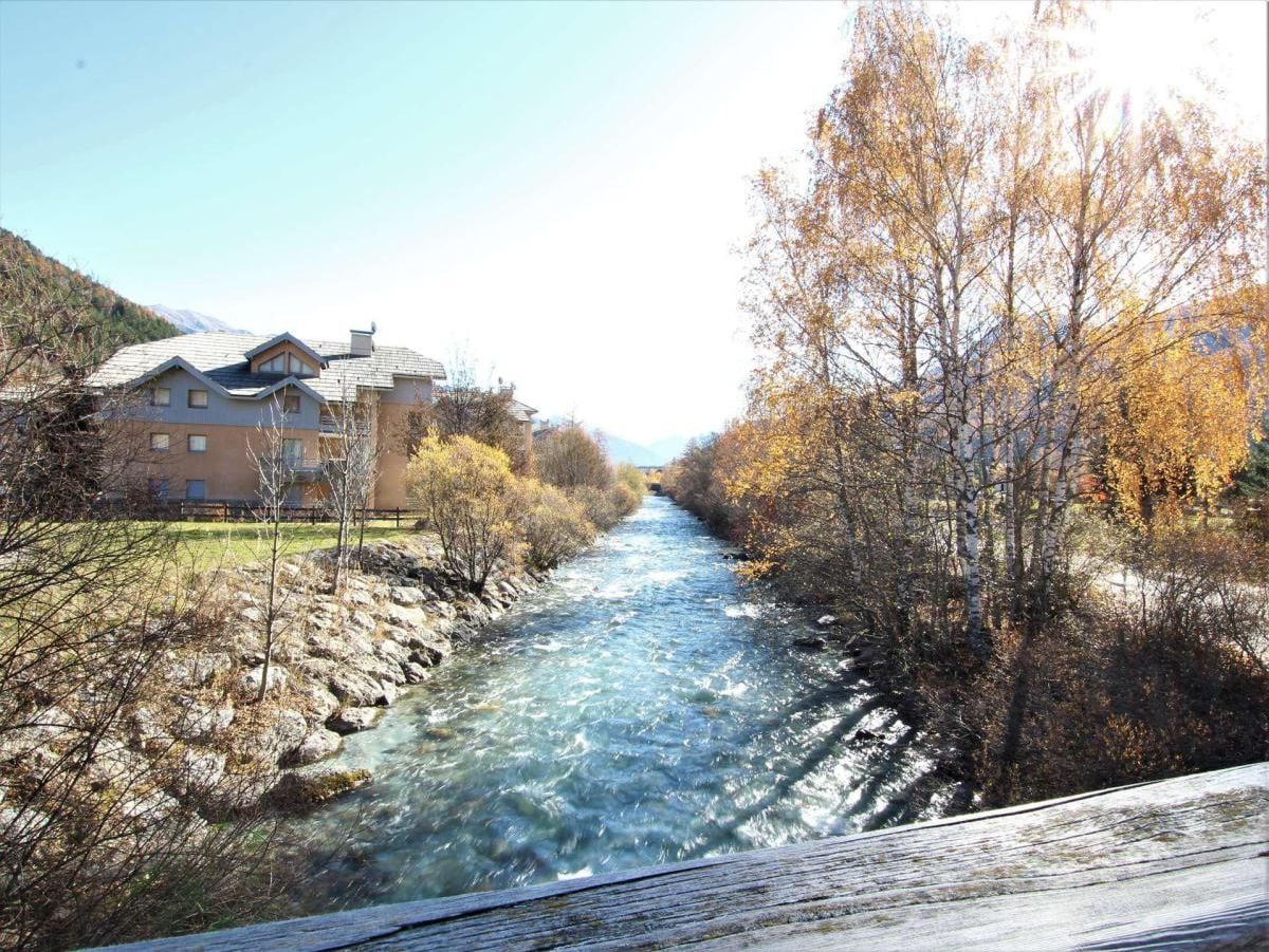 A river flowing through a residential area with houses on one side and trees with autumn foliage on the other.