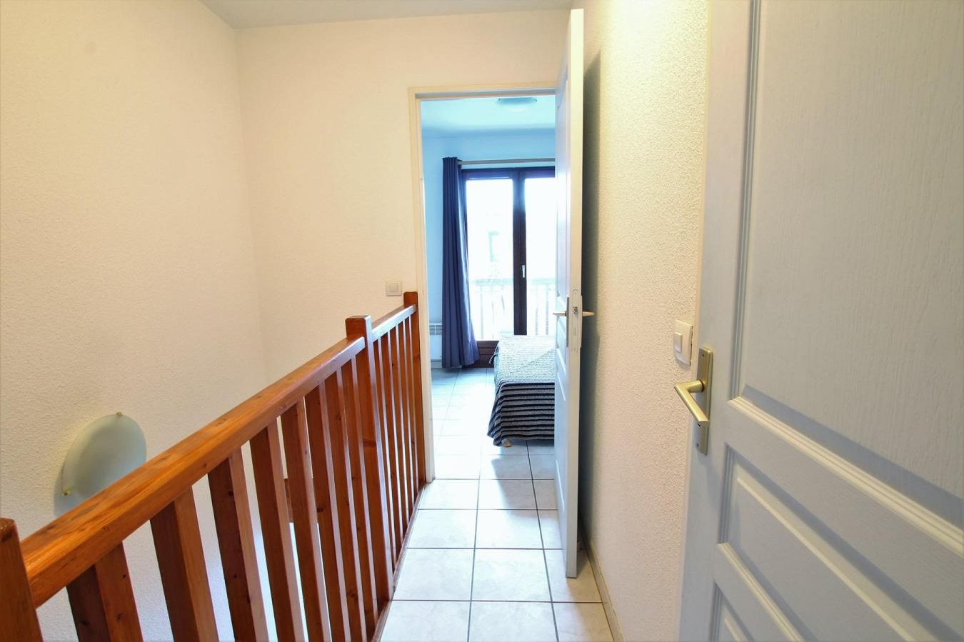 A narrow hallway with a wooden railing, leading to a room with a bed and a window with curtains.