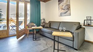 A cozy living room with a grey sofa, a wooden coffee table with two glasses, a patterned chair, and a large window with a view of a snowy landscape. - 200m from ski slopes, Ground floor, terrace, tv, 42m², Les Menuires (Les Menuires)