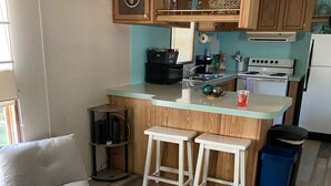 Una cocina con armarios de madera, una encimera blanca y una estufa integrada. Hay dos taburetes blancos y un bote de basura azul.