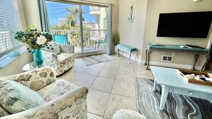 Ein geräumiges Wohnzimmer mit einer Couch, einem Couchtisch und einem Fernseher. Ein Balkon bietet Blick auf Palmen und Gebäude.