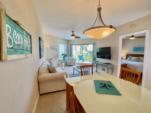 A spacious living area with a sofa, dining table, and chairs. There is a TV mounted on the wall, a ceiling fan, and a sign that reads "Red House".