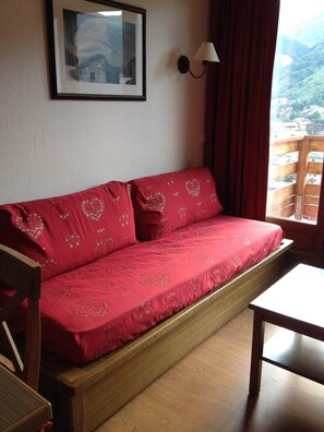 Un canapé rouge orné de motifs en forme de cœur, un tableau encadré accroché au mur, une lampe et une vue sur une région montagneuse, visible par une fenêtre.