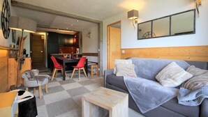 Een gezellige woonkamer met een grijze bank, een houten salontafel en rode stoelen. Er is een eethoek met een tafel en stoelen, en een keuken op de achtergrond.