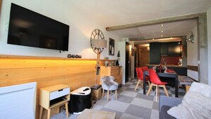Een gezellige woonkamer met een televisie, houten lambrisering, een eethoek met rode stoelen en een keuken die door een open doorgang zichtbaar is.