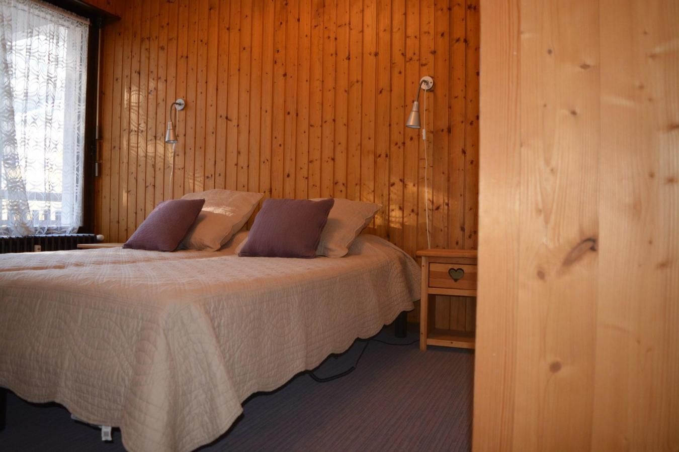 A wooden-paneled bedroom with a bed, two wall-mounted lamps, and a nightstand with a drawer.