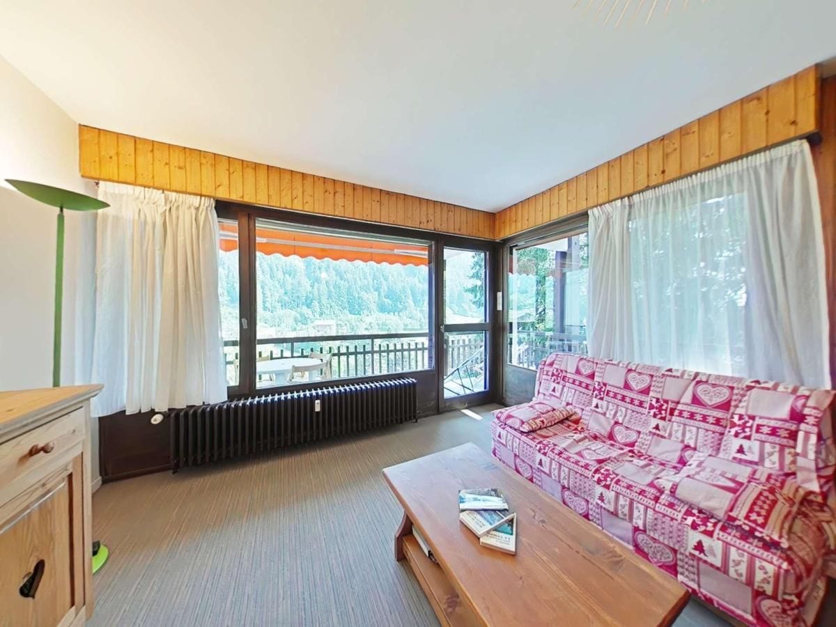 A cozy room with a wooden ceiling, a sofa with a patterned cover, a wooden table with a book, and large windows with curtains.