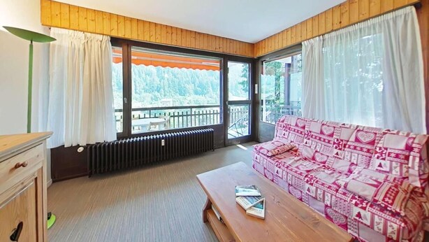 A cozy room with a wooden ceiling, a sofa with a patterned cover, a wooden table with a book, and large windows with curtains. - Resort center, 2nd floor, view mountain, tv, 58m², Le Grand Bornand (Le Grand-Bornand)