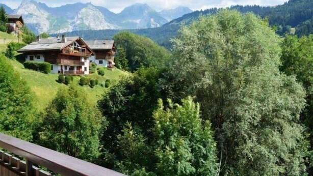 A chalet-style building with a balcony, surrounded by lush greenery and mountains in the background.
