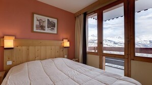 A bedroom with a large bed, wooden headboard, two wall-mounted lamps, a framed picture, and a view of snow-capped mountains through a sliding glass door. - 10m from ski slopes, 5th floor, view mountain, balcony, tv, ski locker, 65m², Montchavin La Plagne (Bellentre)