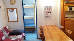 Una habitación pequeña con una mesa y sillas de madera, una cama litera, una kitchenette y un cartel en la pared.