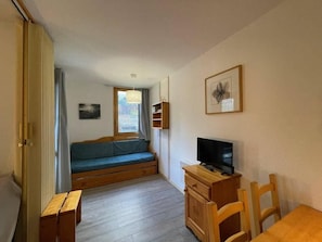 A small room with a wooden bench, a television on a wooden cabinet, a window with curtains, and a framed picture on the wall.