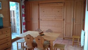 A wooden dining table with six chairs, a small stool, and a bowl on the table. There is a wooden cabinet with a door, a small stool, and a refrigerator. - Ground floor, balcony, ski locker, 35m², Pra Loup (Pra Loup)