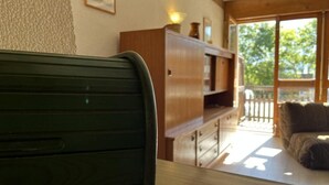 A room with a wooden table, a green chair, a wooden cabinet, and a balcony with a view of trees. - Ground floor, balcony, tv, 30m², Pra Loup (Pra Loup)