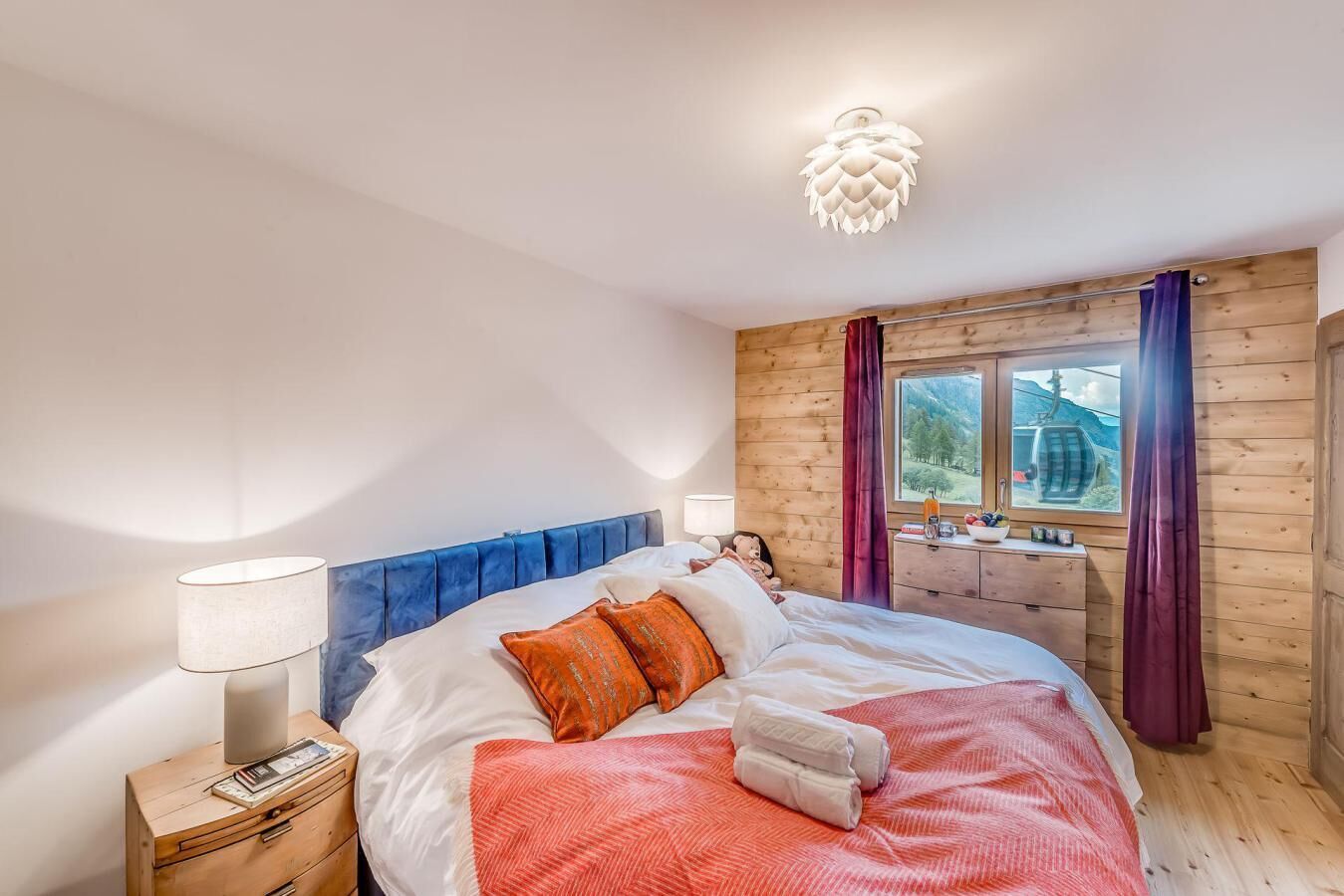 A bedroom with a wooden headboard, a bed with white and orange bedding, a nightstand with a lamp, a window with a view of mountains, and a wooden wall.