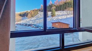 A view through a window of a building with a snowy landscape, a wooden cabin, and parked cars. - Ski-in/ski-out & foot of the slopes, 3rd floor, tv, ski locker, 24m², Les Menuires (Les Menuires)