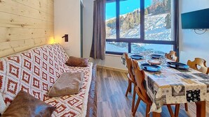 A cozy room with a patterned sofa, a dining table set for two, and a large window offering a view of a snowy landscape. - Ski-in/ski-out & foot of the slopes, 3rd floor, tv, ski locker, 24m², Les Menuires (Les Menuires)