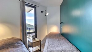 A bedroom with two beds, a window with a view, a bedside table with a lamp, and a teal accent wall.