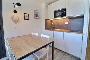 A compact kitchen with a wooden table, white chairs, a black pendant light, a microwave, and a sink.