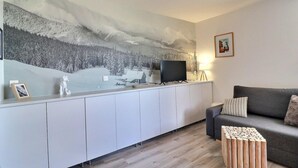 A modern living room with a large wall mural, a flat-screen TV, a grey sofa, and a wooden side table.