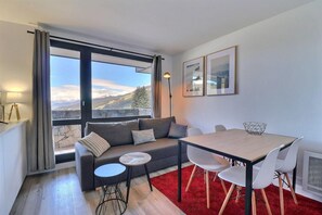A modern living room with a sofa, a small round table, and a dining table with chairs. There are large windows with a view of mountains. - 10m from ski slopes, 2nd floor, balcony, 27m², Méribel-Mottaret (Méribel - Les Allues)