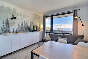 A modern living space with a dining area, a sofa, a TV, and a large window offering a mountain view.