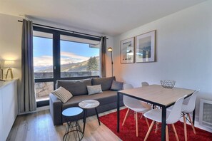 A modern living room with a sofa, a dining table, and chairs. There is a large window with a view of mountains.