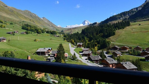 A mountain village with traditional wooden houses, a winding road, and a prominent mountain peak in the background.