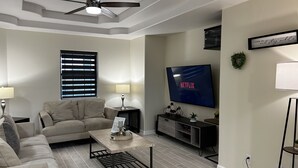 A modern living room with a ceiling fan, a flat-screen TV displaying 'NETFLIX', a sofa, a coffee table, and a side table with a lamp. - Cozy Family-sized Townhome (Pharr)