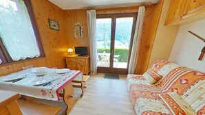 Et hyggeligt værelse med træpaneler, et spisebord dækket med tallerkener og glas, en sofa med mønstrede puder, og en skydedør af glas til et udendørs område.