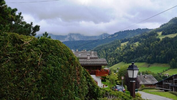 En traditionel schweizisk chalet med skråt tag, omgivet af frodig vegetation og et bjerglandskab.