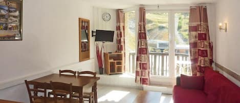A room with a dining table, chairs, a red sofa, a television, and a window with curtains.