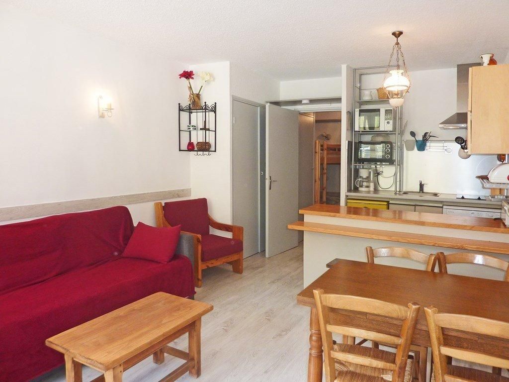 A living room with a red sofa, wooden chairs, a wooden table, and a kitchen area with a countertop and cabinets.