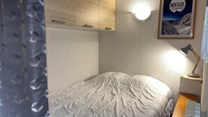 Une chambre à coucher avec un lit, une table de chevet, une lampe, une affiche au mur et un rideau orné d’un motif de feuilles.