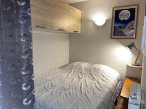Un dormitorio con una cama, una mesita de noche, una lámpara, un cartel en la pared y una cortina con un estampado de hojas.