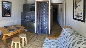 Un espace de vie compact comprenant un coin cuisine, un coin repas avec une table et des tabourets en bois, un canapé et un rideau.