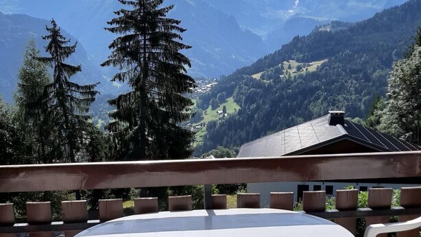 Une terrasse avec une table ronde et des chaises en plastique blanc, offrant une vue sur un paysage montagneux avec des sommets enneigés et une vallée boisée.