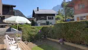 Una casa de madera con techo de metal, un patio con sillas blancas y un paraguas a rayas, y un seto bien cuidado.
