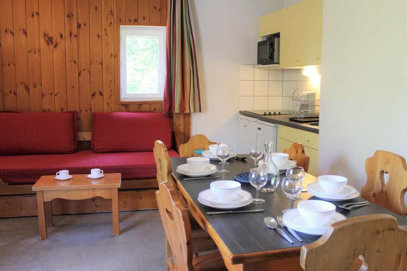 Une pièce chaleureuse avec un plancher en bois, un canapé rouge, une table à manger dressée avec des assiettes, des verres et des couverts, et un coin cuisine équipé d’un four à micro-ondes.